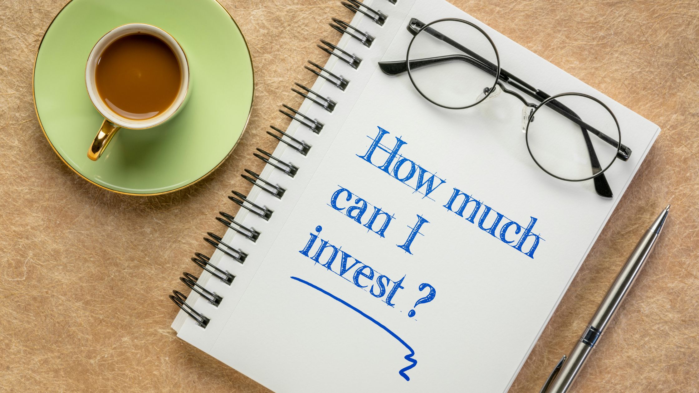 Notepad with "How much can I invest?" written on it, next to a cup of coffee and glasses, illustrating the question "How much should I invest as a beginner?"