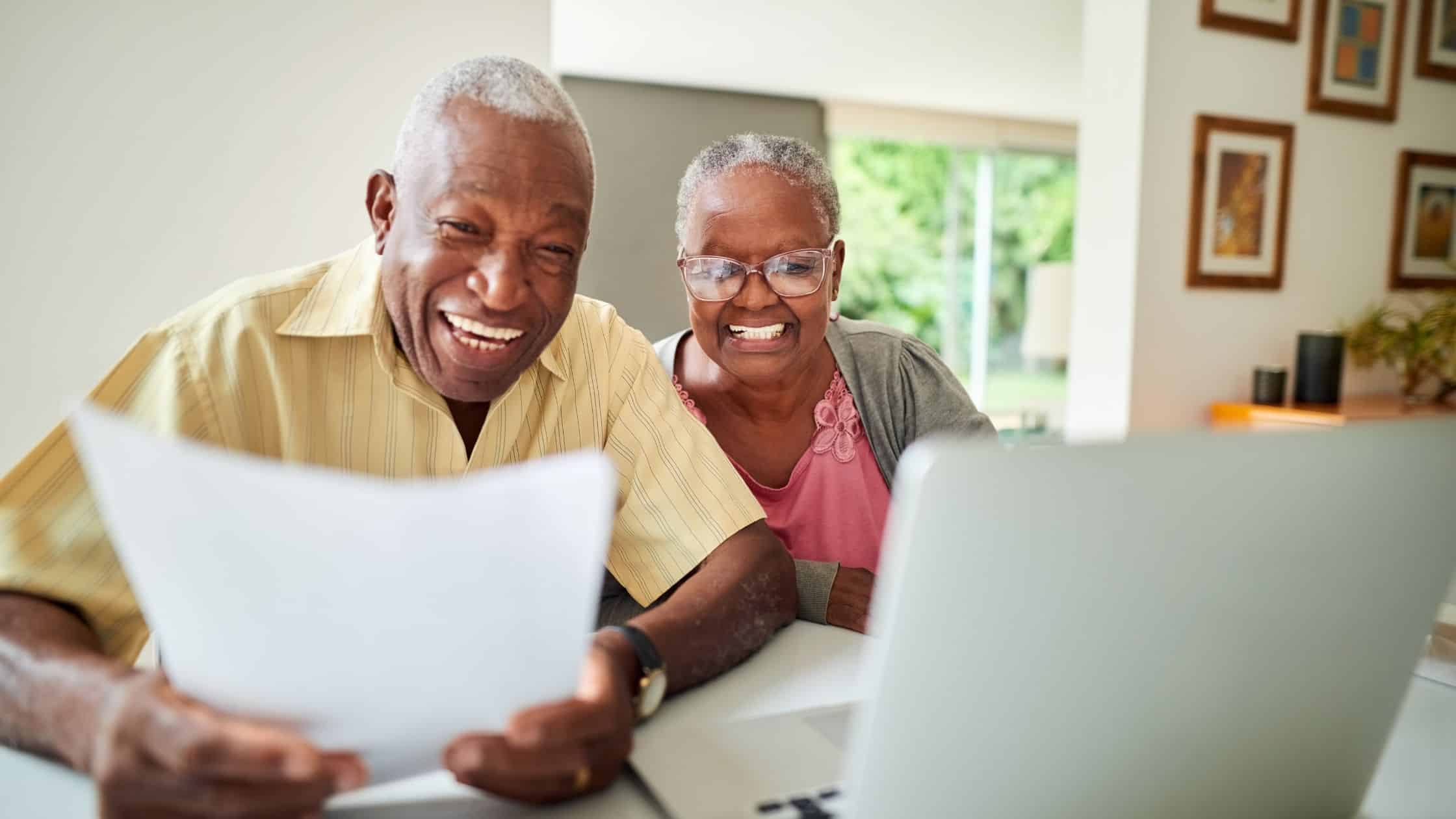 Alt text: A retirement planning fiduciary session: An elderly African American couple smiles happily while reviewing financial documents together on a laptop, likely discussing their retirement strategy with a financial advisor.