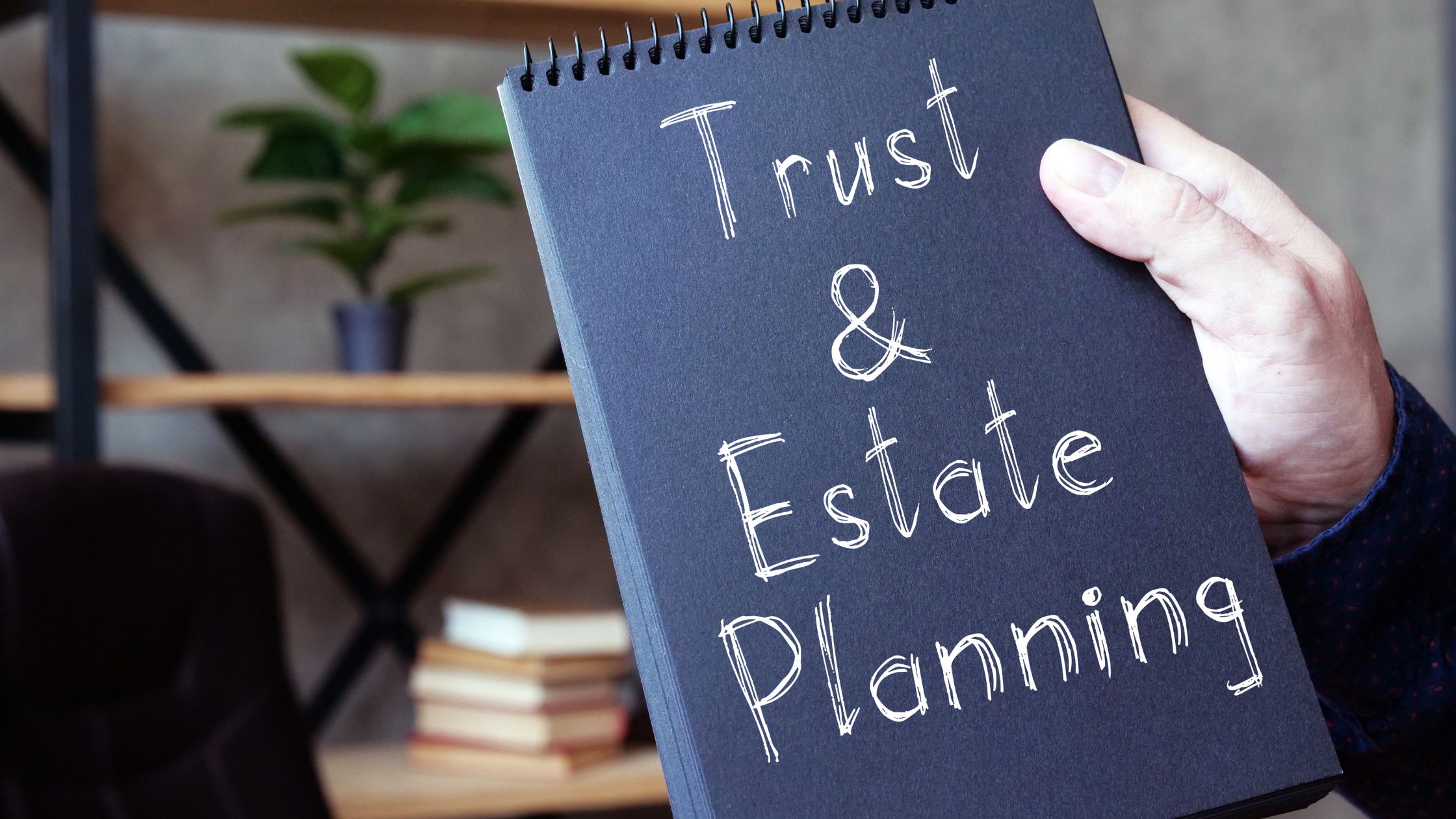 Trust and estate planner's notebook titled "Trust & Estate Planning", held by a hand, with office background.