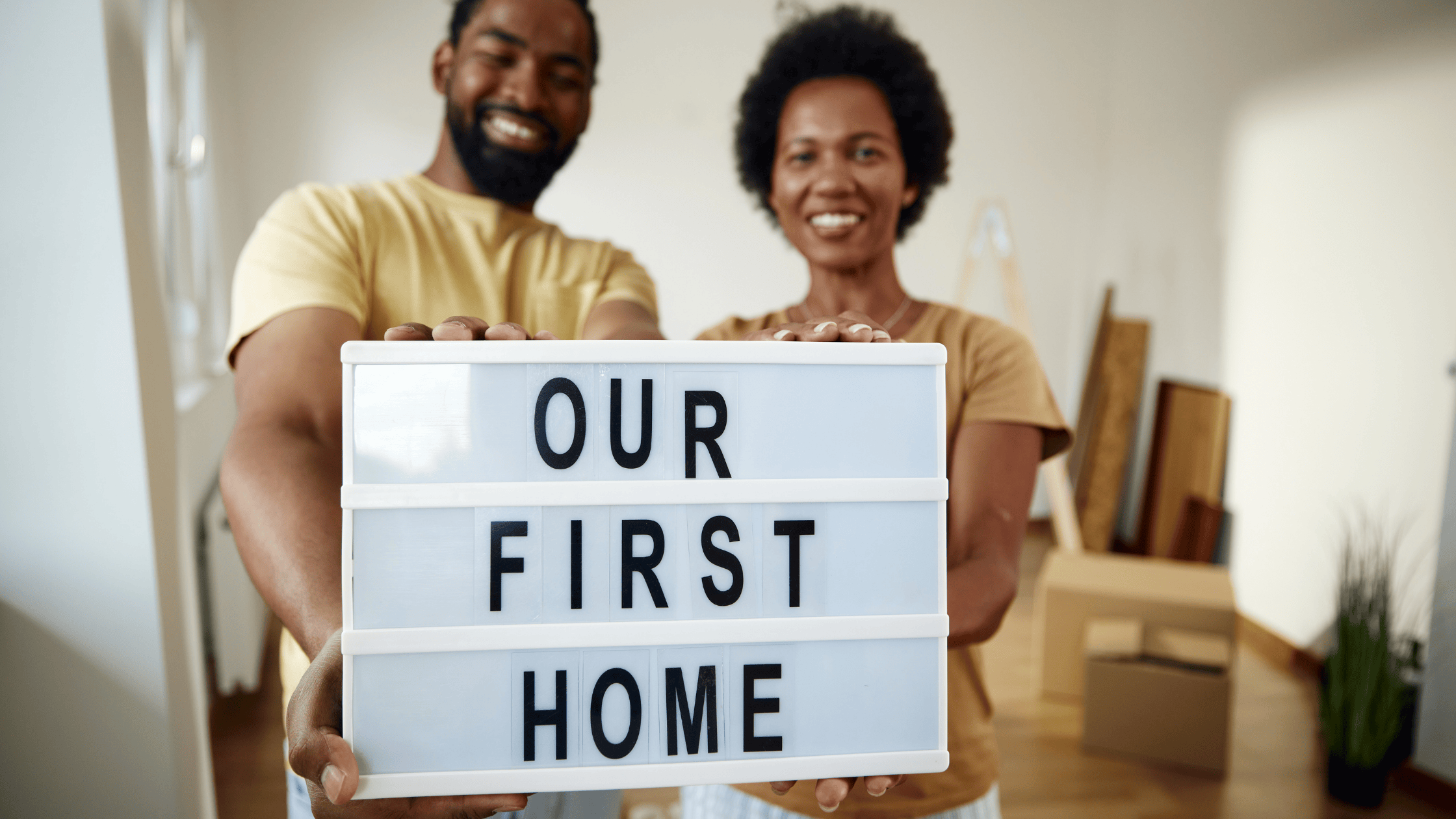 First time home buyer guide: Happy couple holding 'OUR FIRST HOME' sign in their new house with moving boxes in the background.